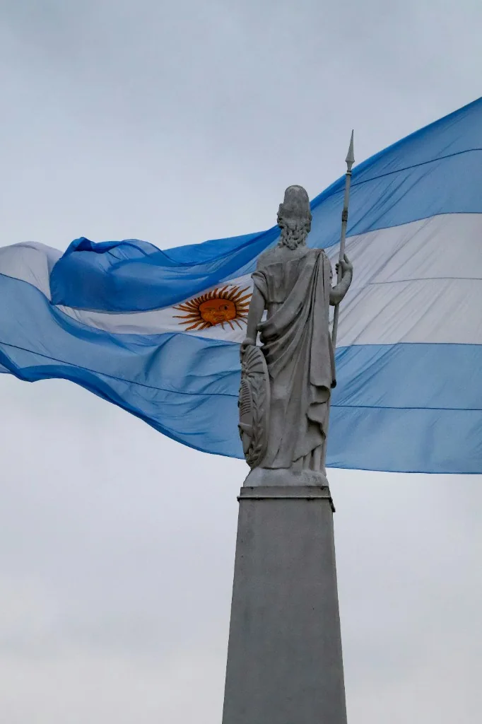 Stone statue of a robed figure holding a spear, with the Argentine flag flying behind it.