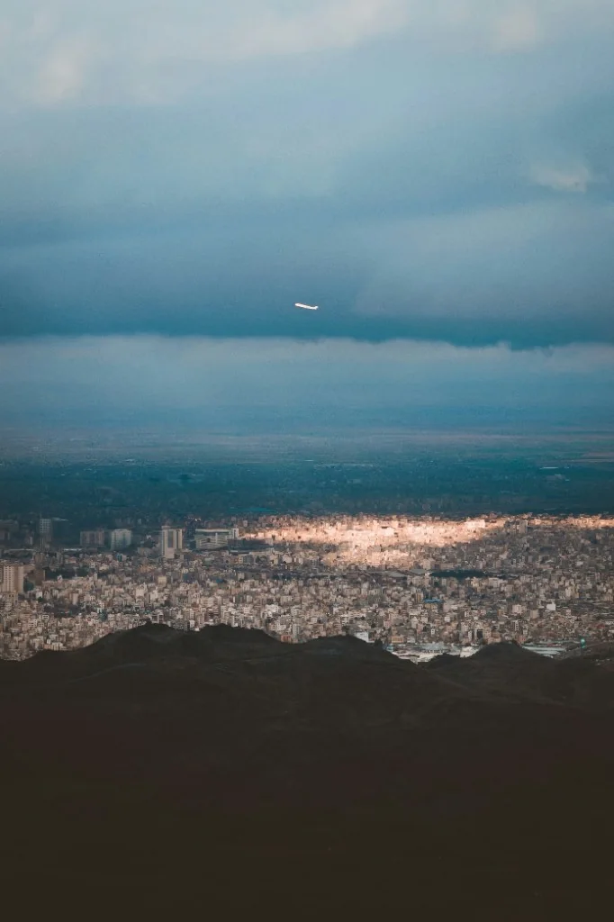 Hillside view over a dense city, with an airplane in a cloudy sky.